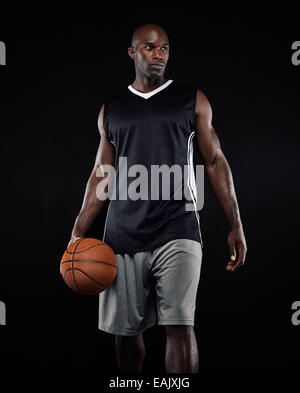 Ritratto di giocatore di basket su sfondo nero. Uomo di afro-americana in azienda di abbigliamento sportivo di una palla da basket che guarda lontano. Foto Stock