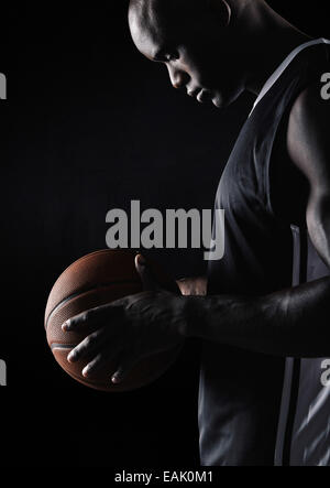 Vista laterale del giovane africano sportivo tenendo la pallacanestro su sfondo nero. Giocatore di basket con sfera. Foto Stock