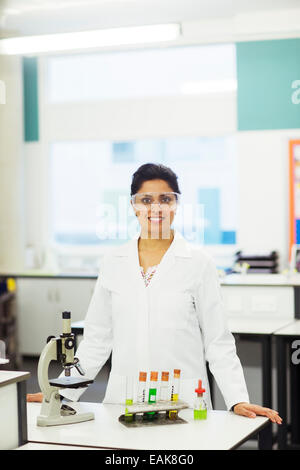 Ritratto di donna sorridente insegnante indossando occhiali protettivi, in piedi dietro la scrivania con microscopio e provette per la prova in rack Foto Stock