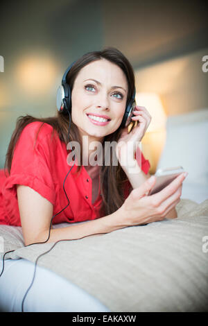 Donna sorridente indossando abito rosso disteso sul letto ascoltando la musica dallo smartphone Foto Stock