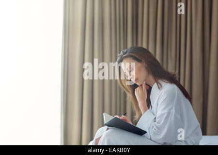 Malinconici donna che indossa accappatoio bianco iscritto nel blocco note in camera da letto Foto Stock