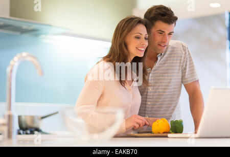 Coppia sorridente tagliare i peperoni e guardando il computer portatile in cucina moderna Foto Stock