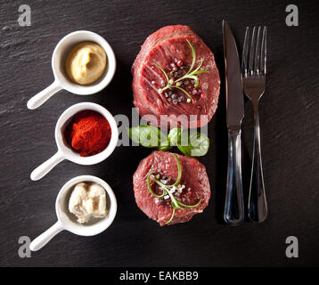 Pezzi di red raw bistecche di carne con erbe aromatiche, servita sulla pietra nera superficie. Scattato dalla vista superiore. Foto Stock