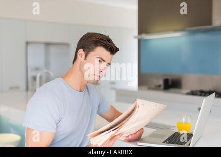 Uomo che legge il giornale in cucina moderna, laptop e succo di arancia sul contatore Foto Stock