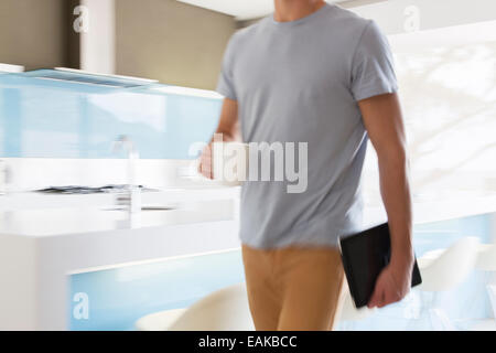 Uomo che porta tazza di caffè e tavoletta digitale a piedi attraverso la cucina moderna Foto Stock