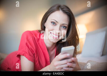 Ritratto di donna sorridente indossando abito rosso sdraiato sul letto con lo smartphone Foto Stock