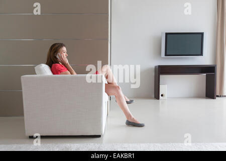 Donna in abito rosso seduto in poltrona e parlando al telefono in soggiorno moderno Foto Stock