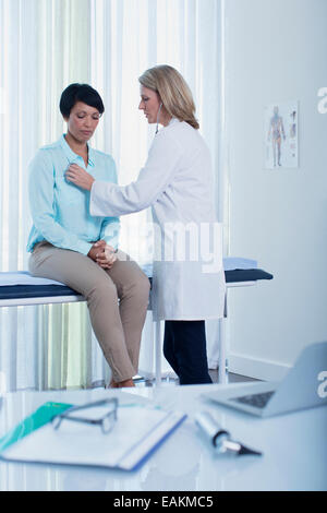 Medico donna esaminando il suo paziente in ufficio, laptop, otoscopio, file e i bicchieri sulla scrivania in primo piano Foto Stock