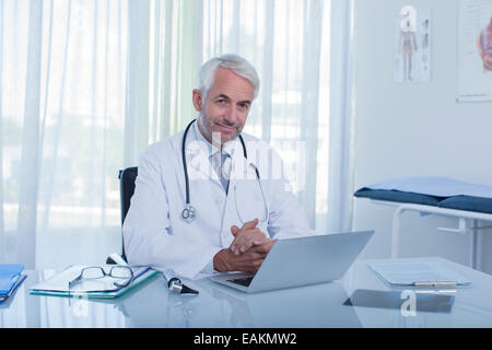 Ritratto di sorridente medico maturo seduto alla scrivania con computer portatile in ufficio Foto Stock