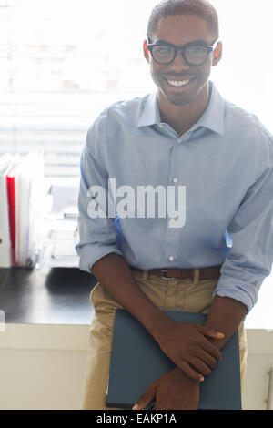 Ritratto di giovane sorridente uomo con gli occhiali e la maglietta blu appoggiato sulla scrivania in ufficio Foto Stock