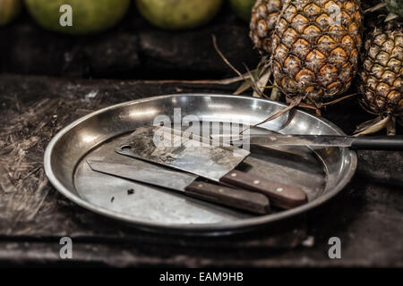 Un chopper un due coltelli su un vassoio di acciaio si trova nei pressi di alcuni ananas Foto Stock