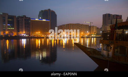 Edifici ad alta contro sky tinte di rosa e le luci della città si riflette nel buio di calme acque del Torrente di Dubai all'alba Foto Stock