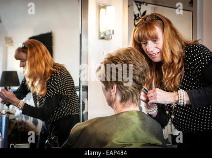 Redheaded parrucchiere con la riflessione nello specchio si appoggia sopra per tagliare i capelli di una signora seduta Foto Stock