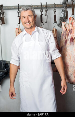 Macellaio in piedi contro la carne appesi in macelleria Foto Stock