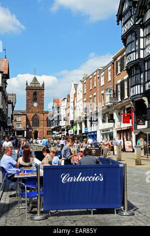 Edifici e negozi lungo Bridge Street con la chiesa di St Peters verso la parte posteriore e un cafe' sul marciapiede in primo piano, Chester, Regno Unito. Foto Stock