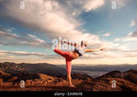 Senior donna fare yoga pone sulla cima della montagna al tramonto con il paesaggio deserto e la valle sottostante. Foto Stock