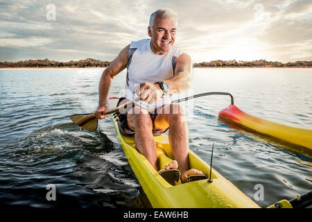 Uomo Senior canoa outrigger canoe nell'oceano al tramonto con isola tropicale dietro di lui. Foto Stock