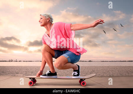 Giovane donna senior su skateboard lungo la spiaggia al tramonto, uccelli in volo in background. Foto Stock