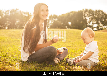 Giovane bella madre che nutre il suo bambino la purea all'aperto alla luce diretta del sole Foto Stock