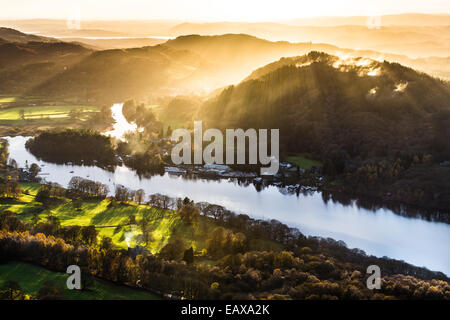 Il fading sole proietta ombre lunghe e raggi attraverso le nebbie sopra l'estremità meridionale del lago di Windermere, Lake District NP Foto Stock