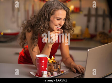 Felice giovane casalinga con spuntini di natale utilizzando laptop in cucina Foto Stock