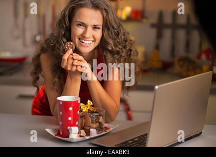 Felice giovane casalinga con spuntini di natale utilizzando laptop in cucina Foto Stock