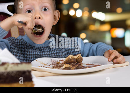 Fame little boy gobbling giù una fetta di gustosa torta al cioccolato con la bocca spalancata per un boccone come egli si siede a un tavolo in un ristorante. Foto Stock