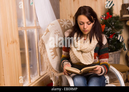 Giovane donna seduta in lettura in una sedia bentwood a fianco di un vetro smerigliato a Natale con un tappeto decorativo con le renne sulla sua spalla. Foto Stock