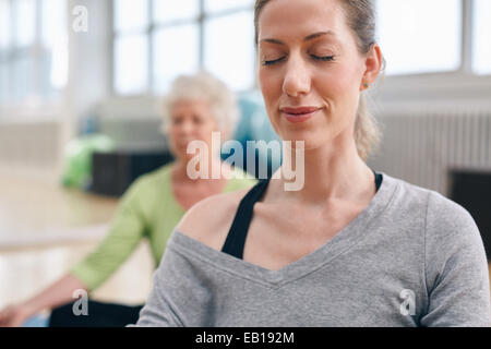 Donna rilassata a praticare yoga in palestra con donna senior in background in meditazione Foto Stock