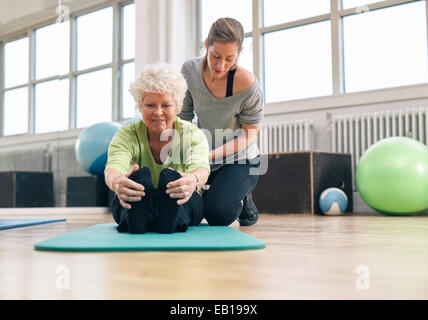 Donna anziana essendo aiutata dal suo istruttore in palestra per esercizio. Senior donna seduta sul tappetino fitness la piegatura in avanti e Foto Stock
