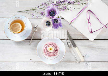 Il tè pomeridiano servito in Cina vintage con tortina con anemone posy matita per notebook e i bicchieri sul tavolo bianco Foto Stock