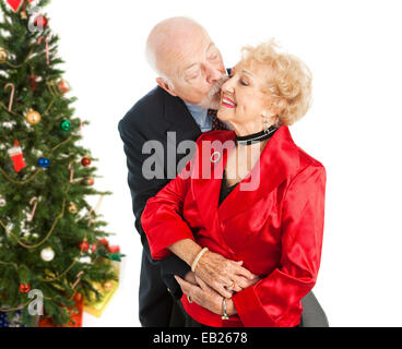Coppia senior vestito per le vacanze. Lui è darle un bacio sulla guancia. Isolato con albero di Natale in background. Foto Stock