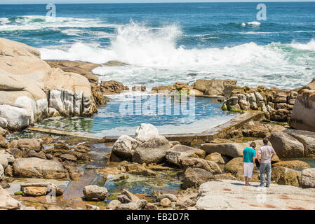 Due persone in piedi sulle rocce al Sea Point, Città del Capo, vicino a un uomo fatto pool di marea, guardando le onde che si infrangono nella roccia Foto Stock