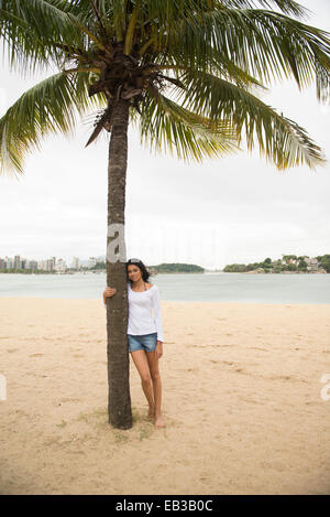 Donna sorridente abbracciando Palm Tree sulla spiaggia tropicale Foto Stock