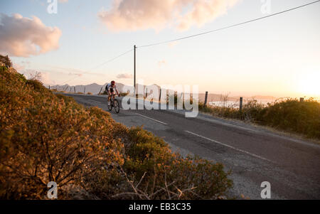 Uomo in bicicletta lungo una strada costiera al tramonto, Corsica, Francia Foto Stock