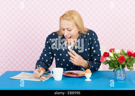 Donna matura rendendo il cruciverba in quotidiani mentre si consuma la colazione Foto Stock