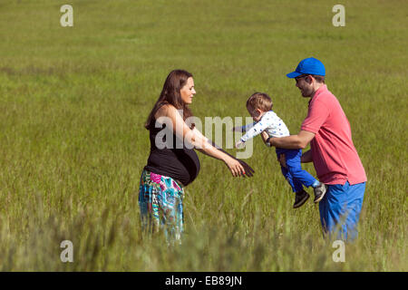Una famiglia felice, una donna incinta con il marito e il bambino trascorrere del tempo insieme e giocare in un prato estivo Foto Stock