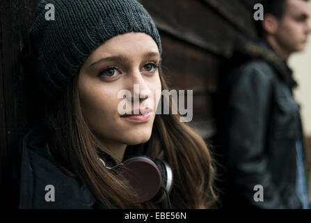 Close-up ritratto di ragazza adolescente all'aperto, indossare un cappello e cuffie intorno al collo con un giovane uomo in background, Germania Foto Stock