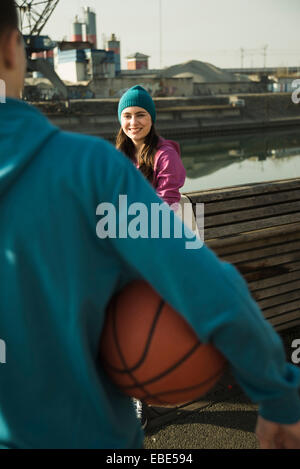 Ragazza adolescente all'aperto indossando toque, sorridente e guardando il ragazzo adolescente tenendo la pallacanestro, zona industriale, Mannheim, Germania Foto Stock