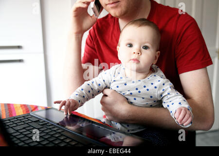 Un padre lavora sul suo computer portatile da casa mentre si tiene il suo otto mesi. Foto Stock