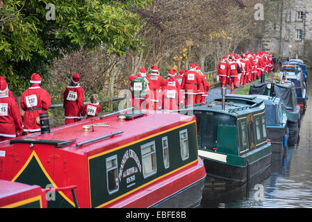 Vista posteriore di persone (uomini, donne e bambini) vestita in rosso e bianco di Babbo Natale abiti passeggiate, jogging, in esecuzione passato canal barche & prendendo parte alla grande Skipton Santa Fun Run, una raccolta di fondi annuale gara di beneficenza organizzata dal Rotary Club - Molle Ramo di Leeds Liverpool Canal alzaia, Skipton Town Center, North Yorkshire, Inghilterra, Regno Unito. Foto Stock