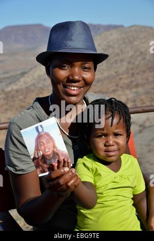 Moderna donna Himba tenendo il suo bambino con una foto di sé indossando vestiti tradizionali, Orupembe, Kaokoland, Kunene, Namibia Foto Stock