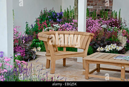 Un giardino contemporaneo con area terrazzata e elegante arredamento di legno circondato da colorati confini di fioritura Foto Stock
