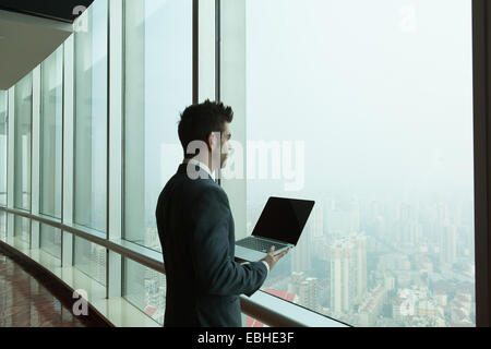 Giovane uomo d affari con computer portatile guardando fuori del grattacielo Finestra di ufficio, Shanghai, Cina Foto Stock