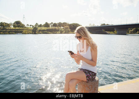 Giovane donna seduta al Riverside texting sullo smartphone, l'Isola del Danubio, Vienna, Austria Foto Stock