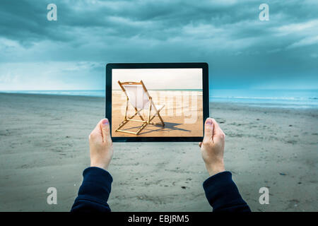 Donna matura sulla spiaggia nuvoloso, tenendo tavoletta digitale mostra sunny beach scena, concentrarsi sulle mani e il computer portatile Foto Stock