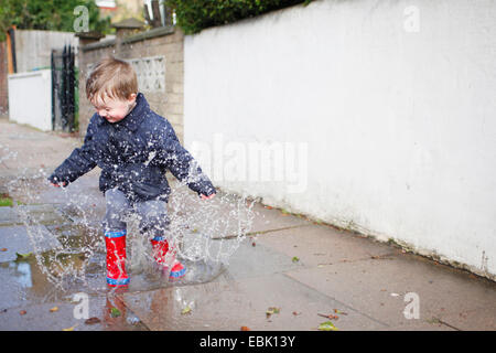 Bimbi maschio in rosso stivali di gomma spruzzi in una pozza sul marciapiede Foto Stock