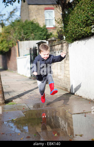 Bimbi maschio in rosso stivali di gomma un salto nella pozza sul marciapiede Foto Stock