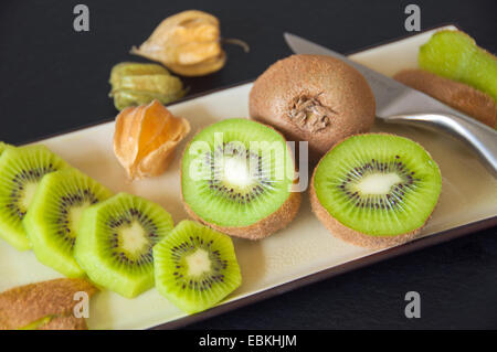 Kiwi e physalis servita in una piastra rettangolare, su uno sfondo nero. (Dimezzato e fette di kiwi). Foto Stock