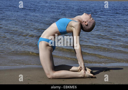 Bald donna in posizione di yoga ustrasana, o cammello, su una spiaggia. Foto Stock